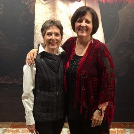 Two women smiling, arms around each other, pose in front of a painting. One in a black vest, the other in a red shawl.