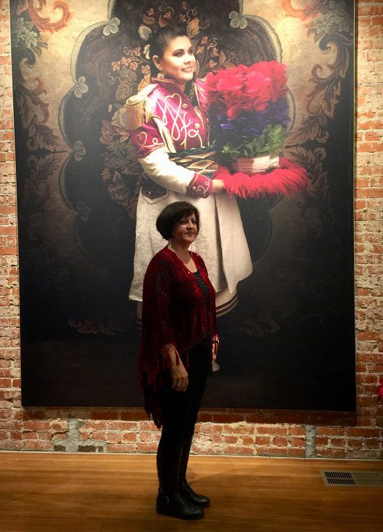 Woman stands before a large painting of a person in a colorful military-style uniform.