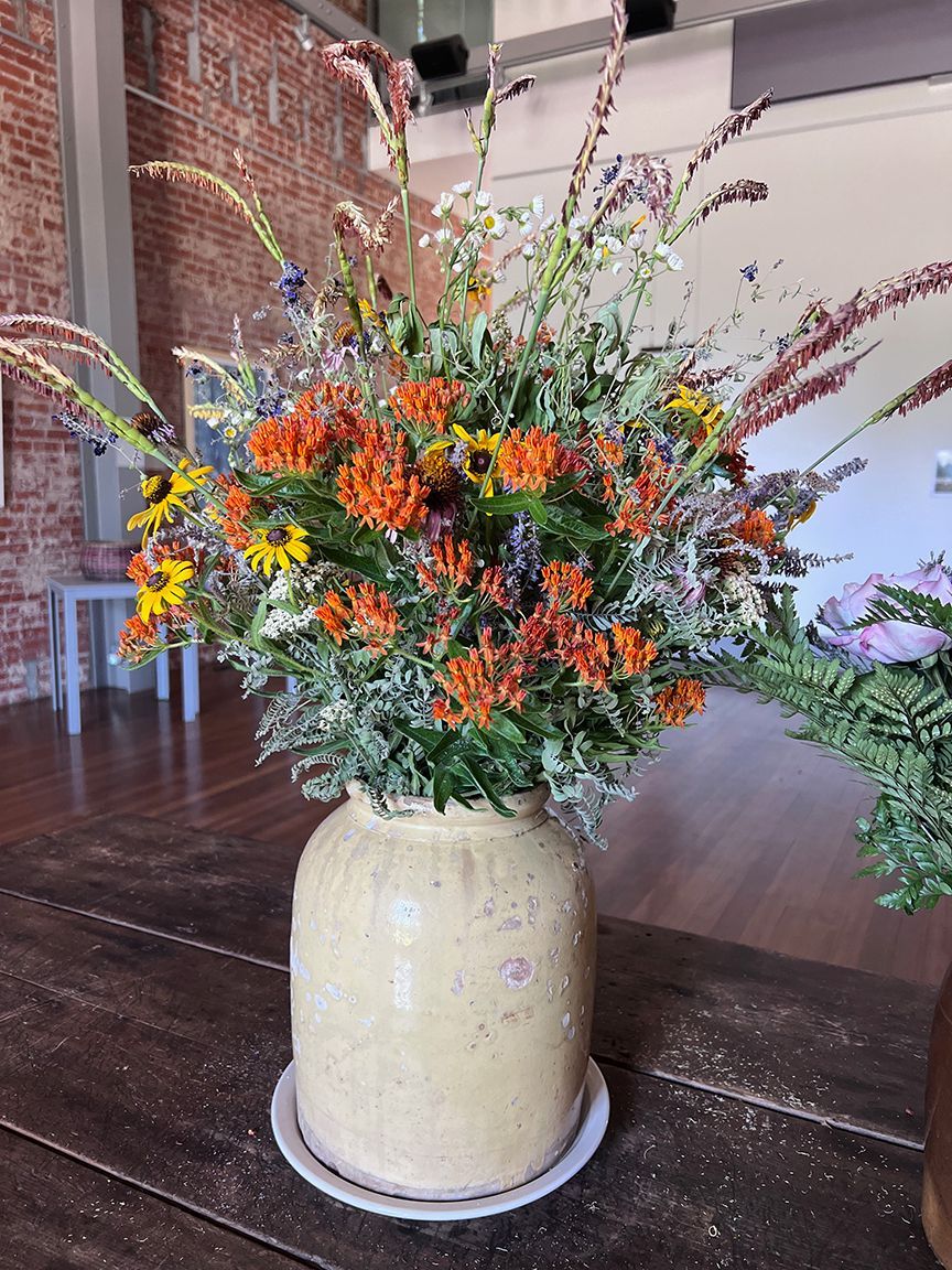 A large bouquet of orange and yellow wildflowers in a cream-colored vase on a wooden table.