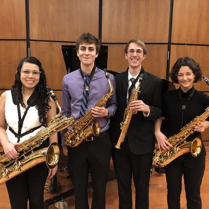 Four young people, each holding a saxophone, smiling. Indoors, music room setting.