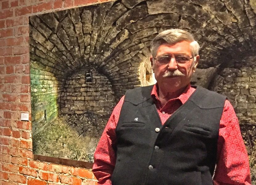 A man with a mustache is standing in front of a painting on a brick wall.