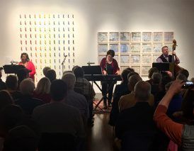 A jazz trio performing in an art gallery, with a seated audience in front.