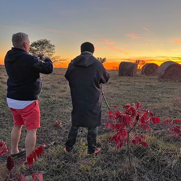 Two people photographing a sunset over a field with hay bales and red plants.