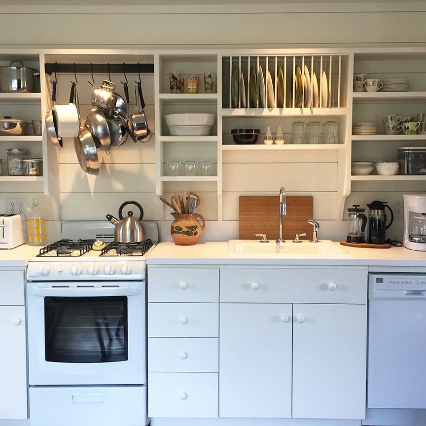 White kitchen with open shelves, pots hanging above a stove, and a plate rack.