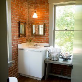 Brick-walled bathroom with white sink, small mirrors, window, and bench holding towels.