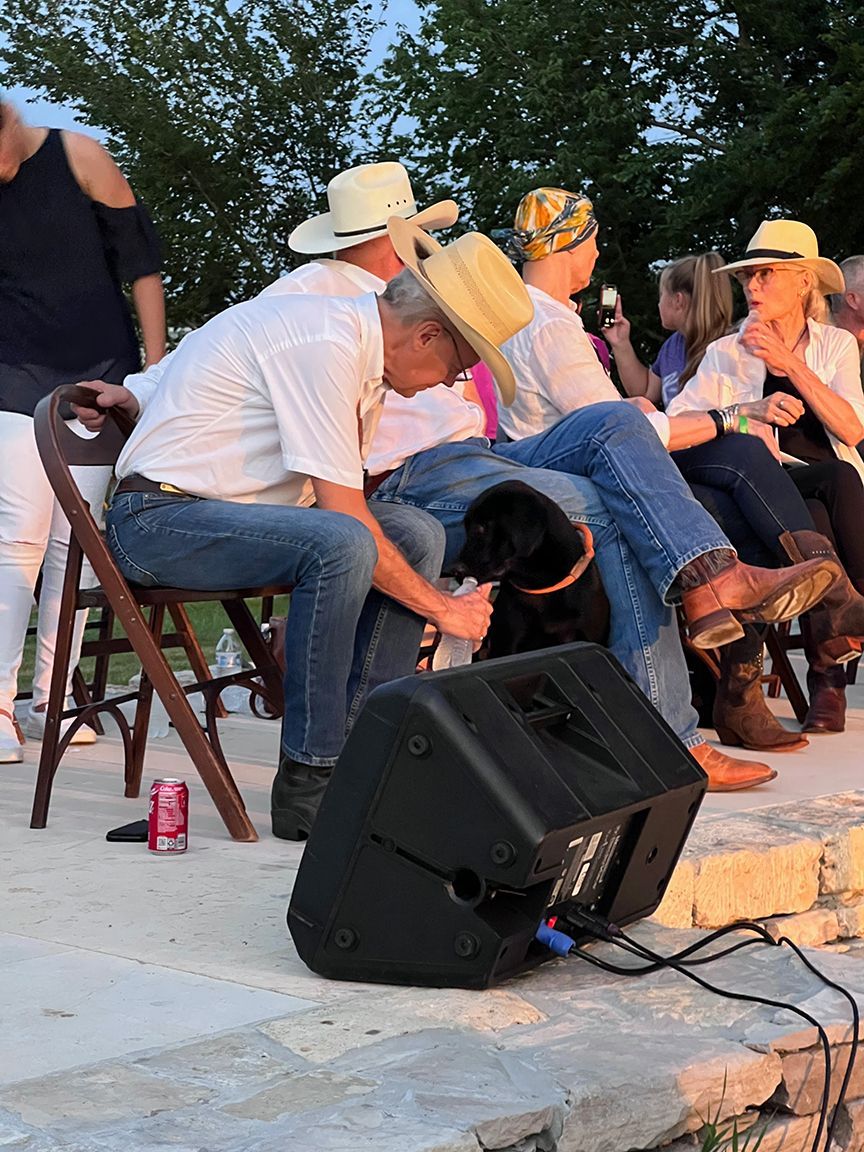 People in cowboy hats and jeans sit outdoors, petting a black dog near a speaker and a stone wall.