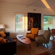 Living room with modern furniture, windows, artwork, and a rug. Sunlight streams through a window.
