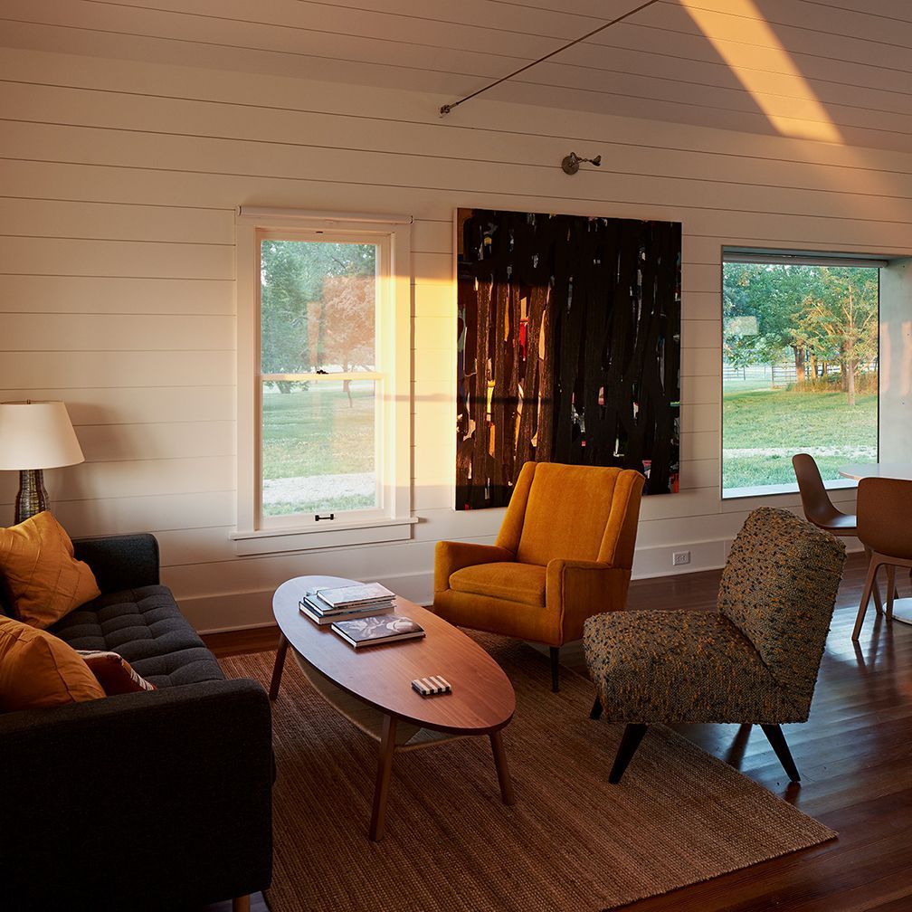 Cozy living room with black sofa, yellow chair, patterned chair, and large artwork; sunlight streams through windows.