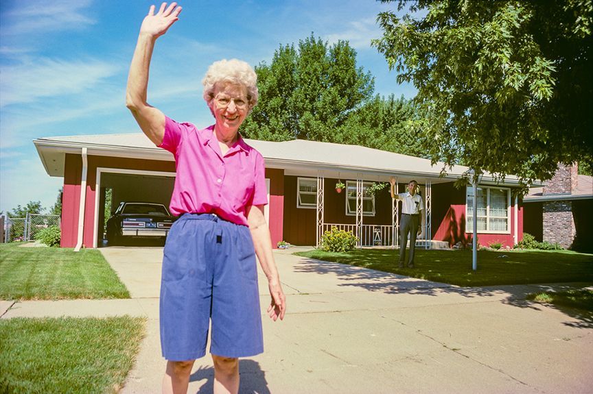 Woman in pink shirt and blue shorts waves from driveway in front of a red house.