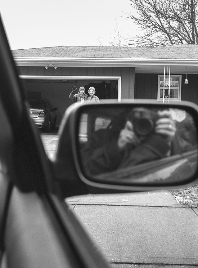 A person takes a selfie in a car; two people wave from a house's garage.