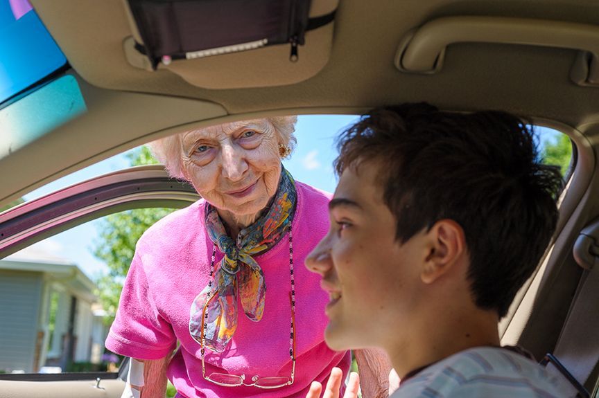 An older woman in pink shirt smiles as she leans into a car, talking to a young man sitting inside.