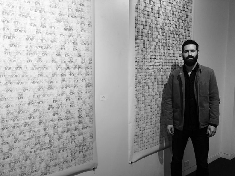 A man with a beard is standing in front of two paintings on a wall.