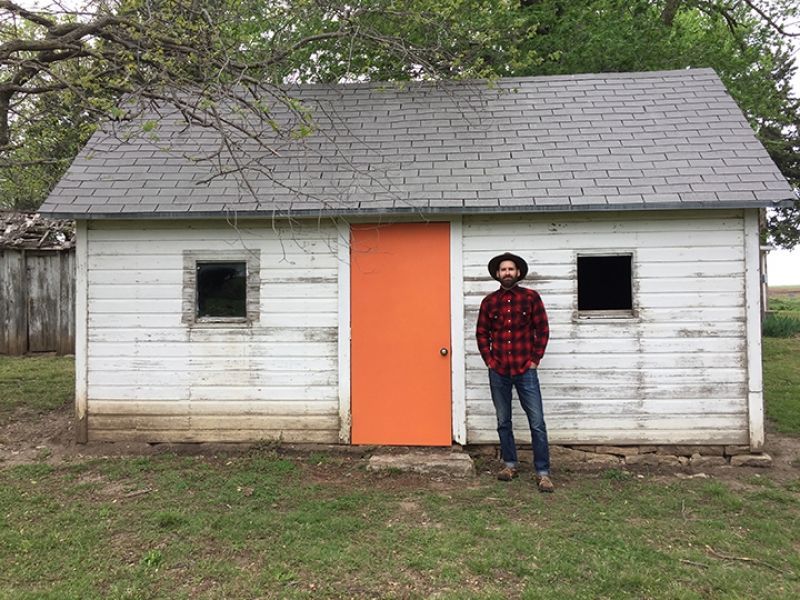 A man in a plaid shirt is standing in front of a small white house with an orange door.
