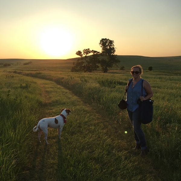 A woman and a dog are standing in a field at sunset