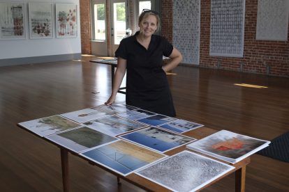 Woman smiles in art gallery, standing by table displaying abstract art, brick wall in background.