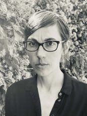 Woman with glasses looks directly at the camera, dark shirt, in front of a leafy background.