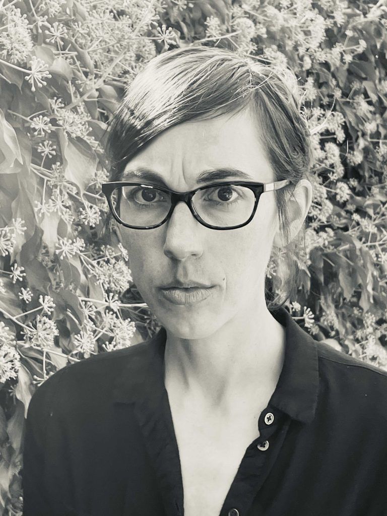 Woman with glasses, short hair, serious expression, wearing a dark shirt, in front of leafy vines.