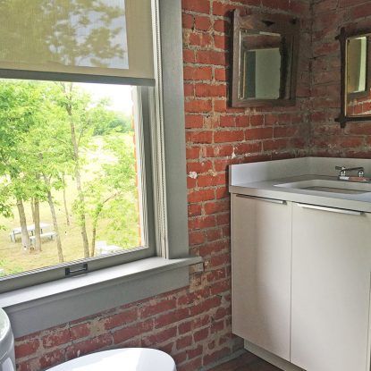 Bathroom with brick wall, window overlooking trees, white vanity, and two mirrors.