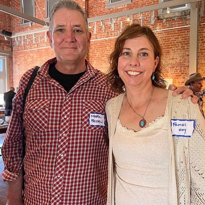 Man and woman smiling, indoors. Man wearing plaid shirt, woman with sweater, both wearing name tags.