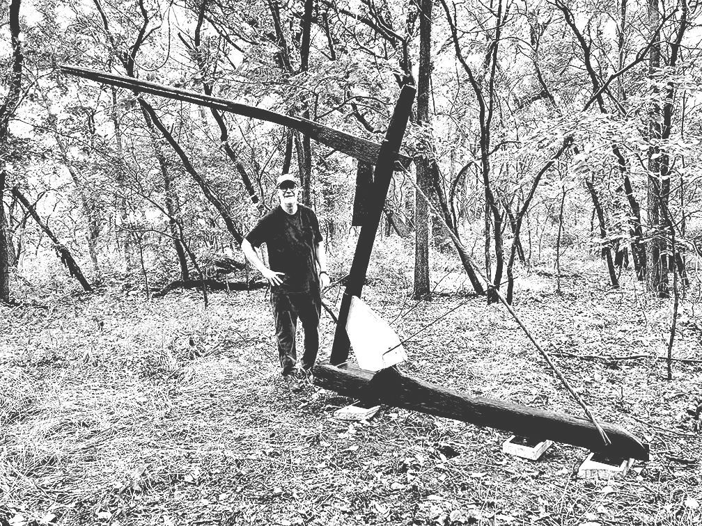 Man stands next to large, fallen wooden structure in a forest.