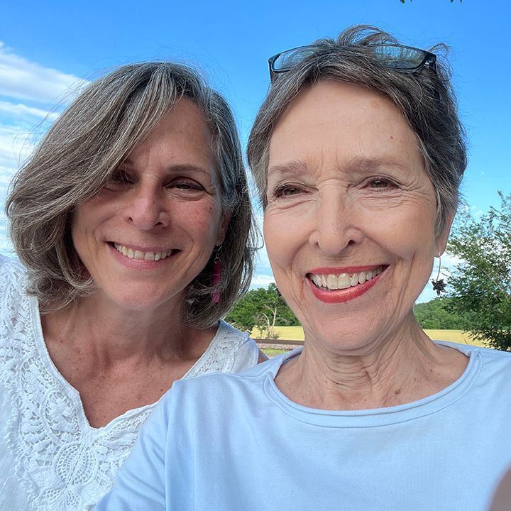 Two women smiling outdoors. One in white lace, the other in light blue. Sunny day, blue sky.