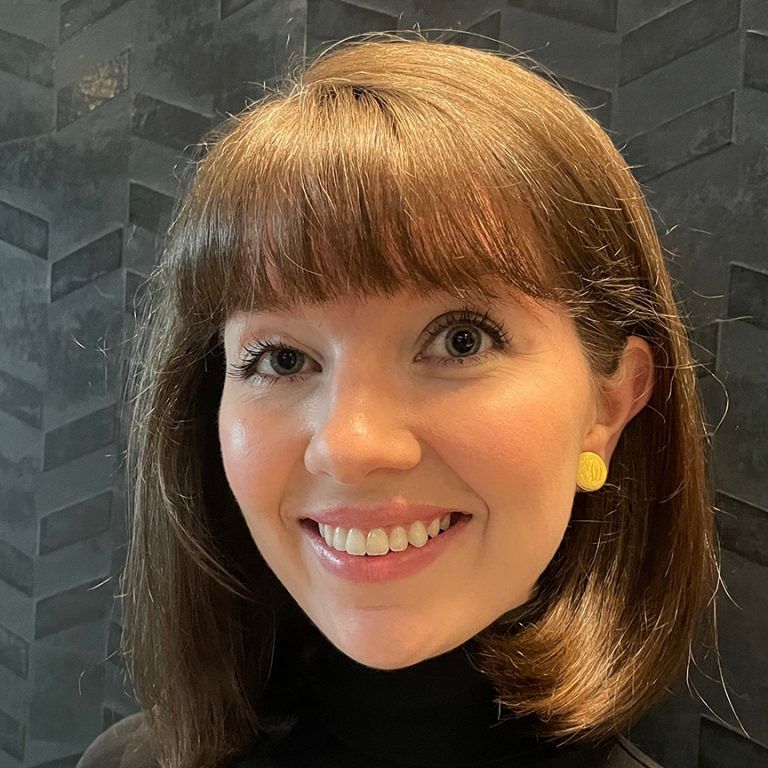 Woman with brown hair and bangs smiles, wearing gold earrings and black turtleneck.