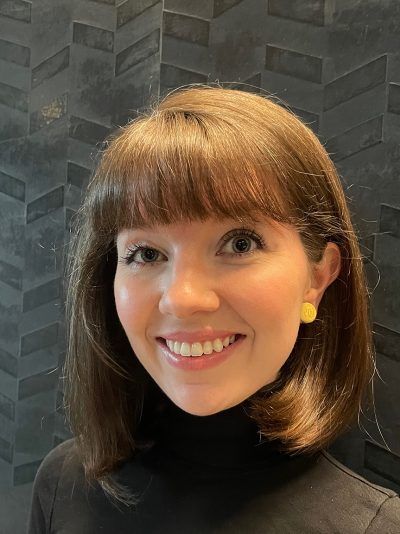 Woman with brown hair and bangs smiles in front of a textured gray wall. She wears a black turtleneck and gold earrings.