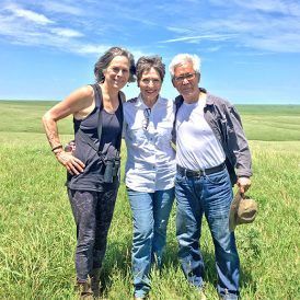 Three people stand in a grassy field on a sunny day. They are smiling and posing for a photo.