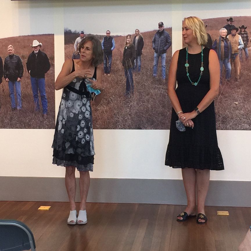 Two women standing in front of a photo display; one speaks with hand gestures, other observes. Indoors.