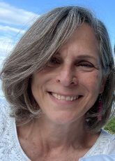 Woman with gray hair smiles outdoors, wearing a white shirt; blue sky background.