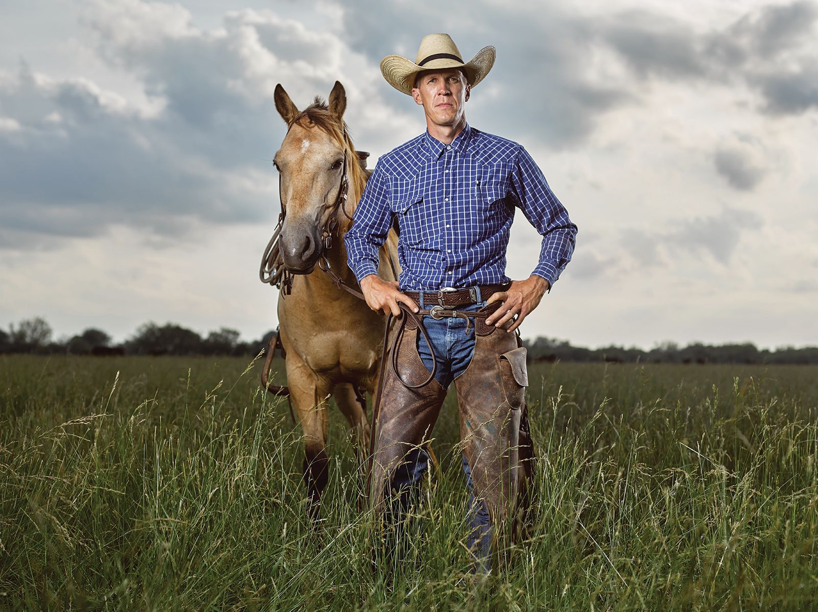 Cowboy standing in tall grass with horse; blue shirt, chaps, cowboy hat; outdoors.