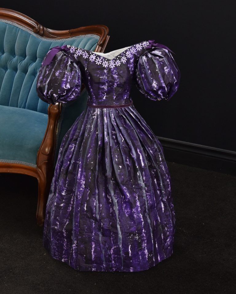 Purple, patterned Victorian-era dress with puffed sleeves; displayed near a blue velvet chair.