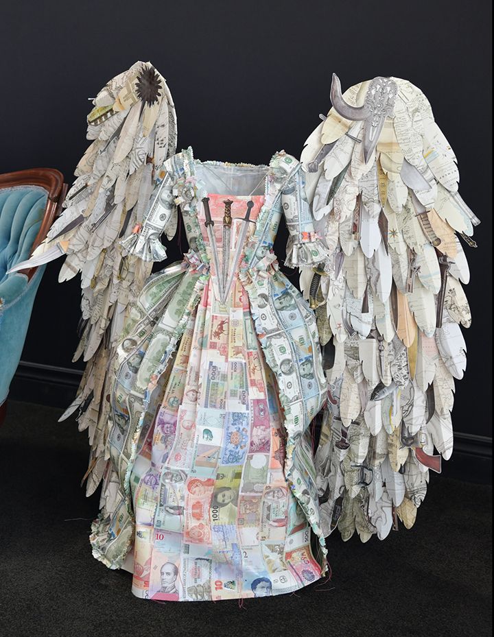 A headless figure in a dress made of currency, with large wings made of book pages.