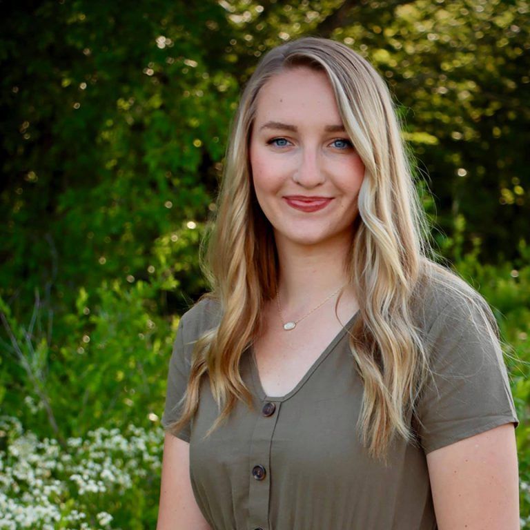 Blonde woman in green dress smiles outdoors in front of leafy foliage.