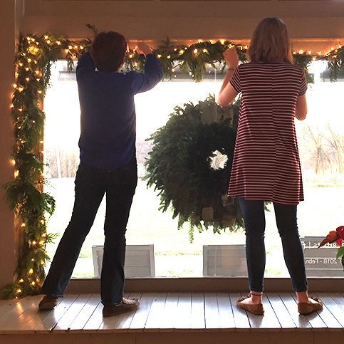 Two people decorating with garland and a wreath in front of a window.