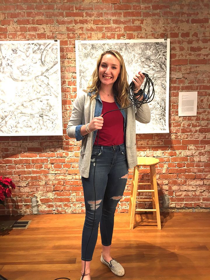 Woman in jeans and blazer, holding cords, smiling in front of art and a brick wall.