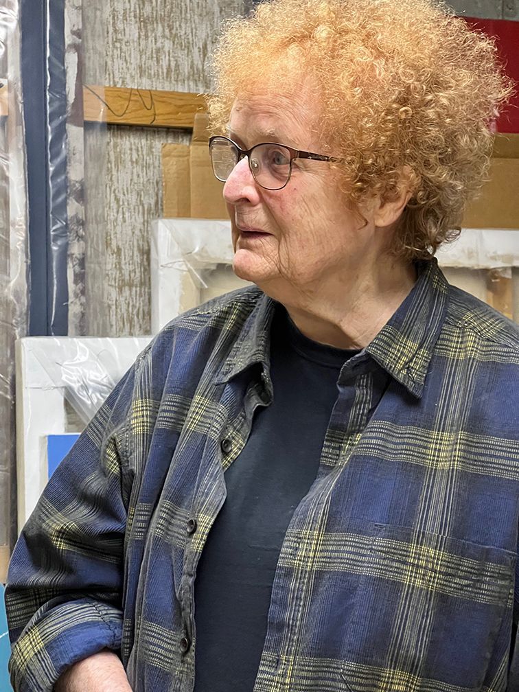 Woman with curly blonde hair, glasses, and a blue plaid shirt, looking to the side.