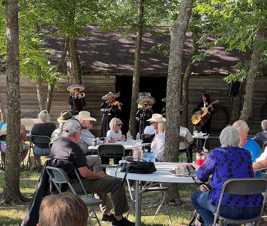 Mariachi band performing outdoors for seated audience near a rustic wooden building.