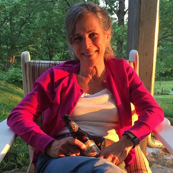 Woman in pink cardigan sits in rocking chair, holding a beer bottle, smiling outdoors.