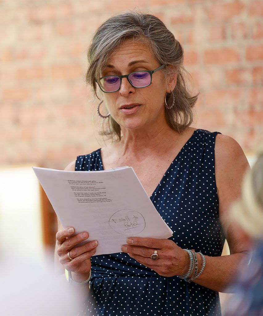 Woman with glasses reading from a paper, wearing a navy polka dot dress. Brick wall background.