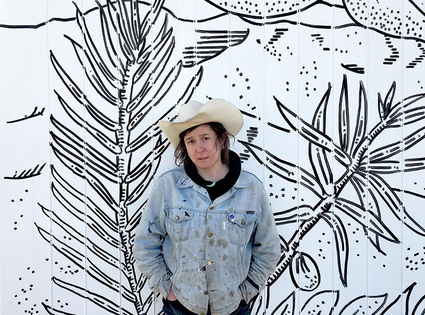 A man wearing a cowboy hat and a denim jacket is standing in front of a wall with drawings on it