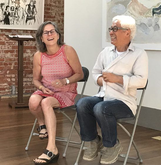 Woman and man laughing while seated on chairs in an art gallery, red brick wall, art in background.