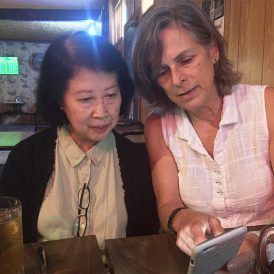 Two women looking at a phone. One Asian woman, one white woman; both are inside at a table, possibly a restaurant.