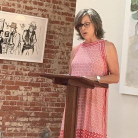 Woman in red patterned dress speaking at podium, brick wall backdrop with artwork.