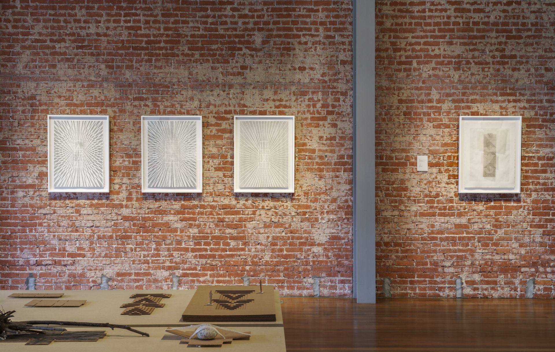Four framed artworks on a brick wall, alongside a light-colored metal support beam. Wooden floor, objects on a table.