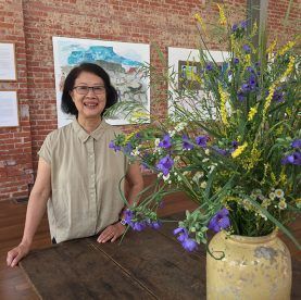 Woman in tan shirt stands by a vase of flowers, brick wall background, art gallery.