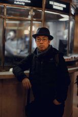 Man in a fedora, glasses, and dark jacket leans against a ticket counter.