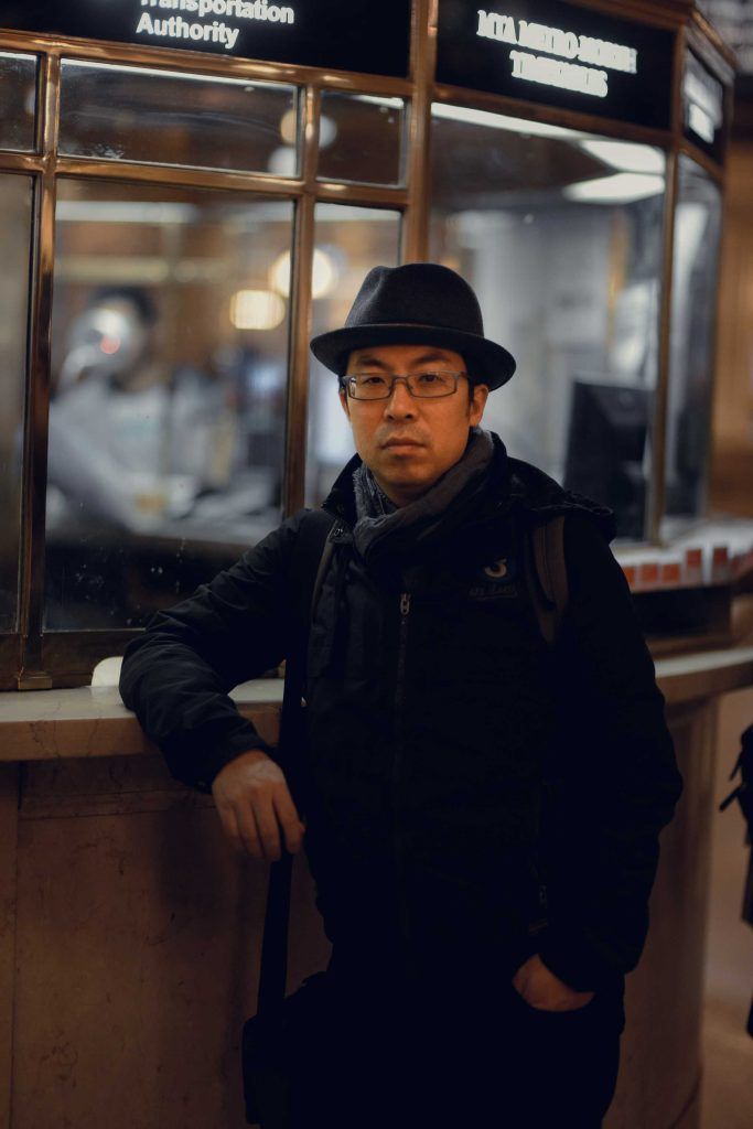 Man wearing a hat and glasses, leaning against a ticket window in a train station.