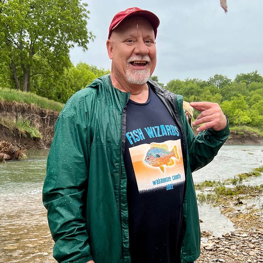 Man in red cap and green jacket smiling by a river, wearing a
