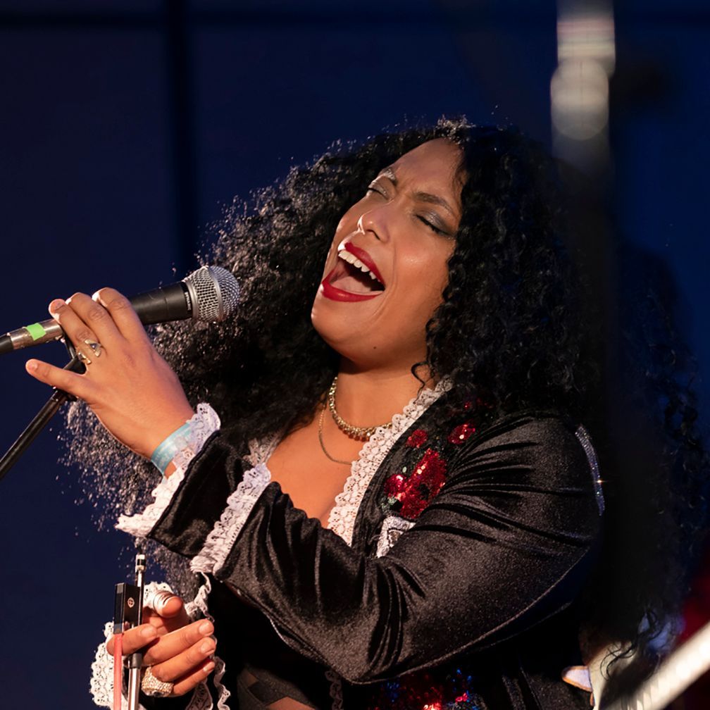 Woman with curly black hair, singing into a microphone, wearing a black jacket with red accents.